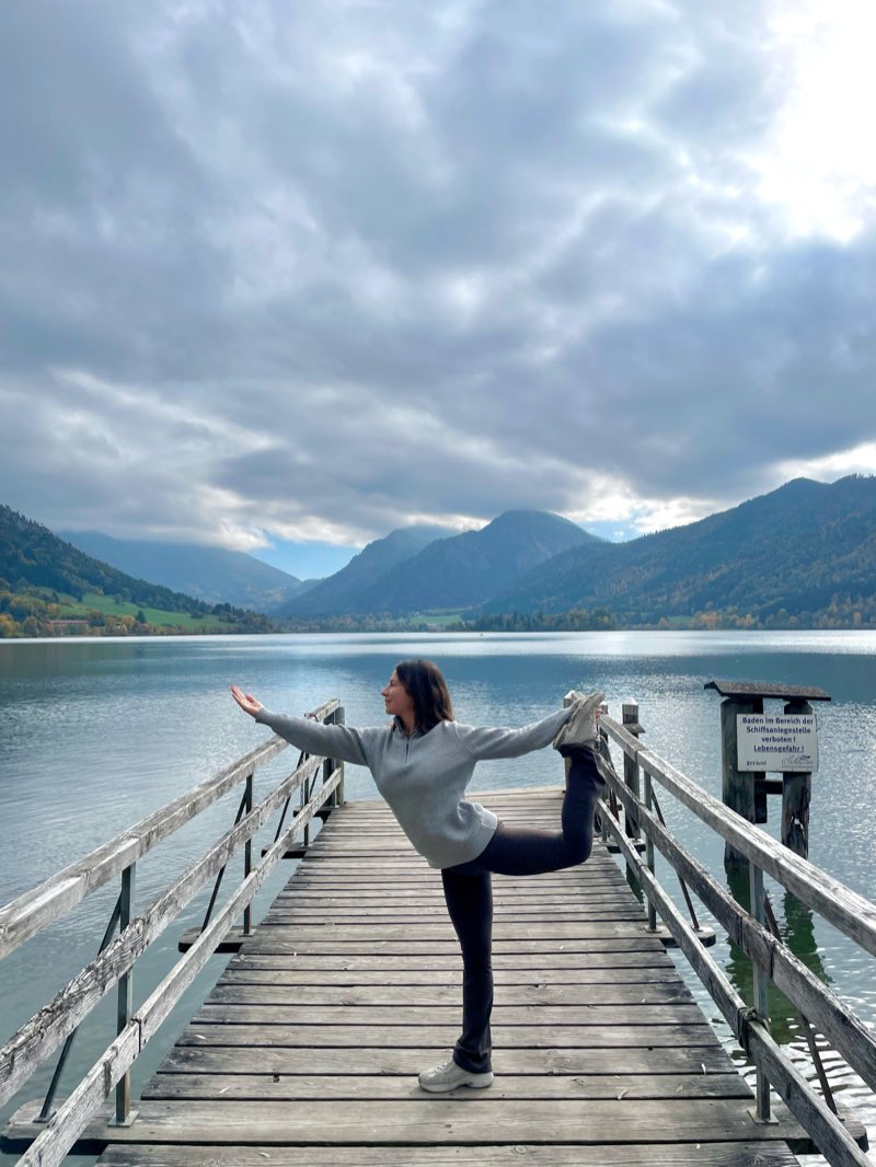 Selin in dancer pose on a wooden jetty over a still mountain lake, under low autumn cloud.