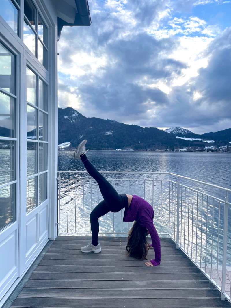 Selin in a standing split on a lakeside deck, snow-covered mountains across the water.